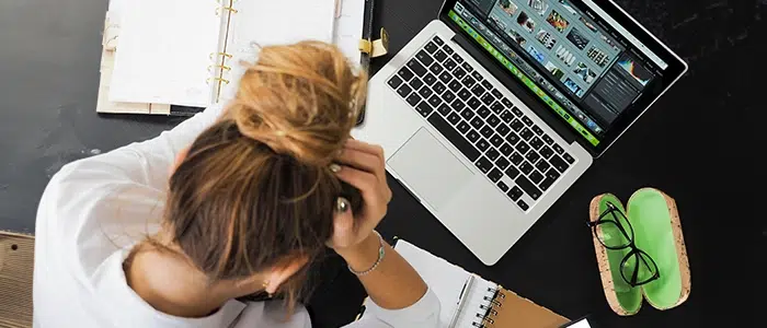 Persona con gesto de tristeza sentada frente a un escritorio donde hay un computador, lentes, celular y libretas.