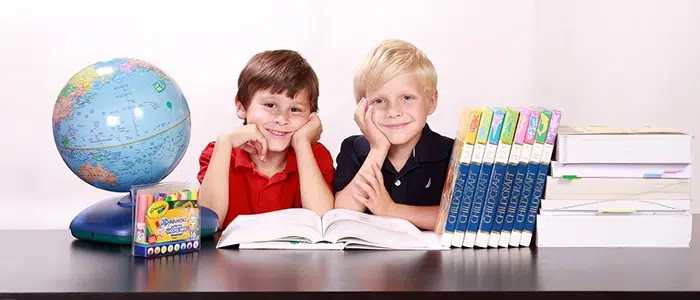 Dos niños sentados frente a un escritorio con libros, lápices de colores y un mapamundi.