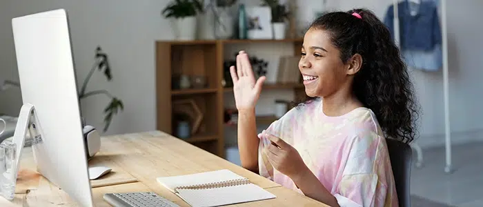 Niña sentada frente a un computador con la mano levantada mientras sonríe.