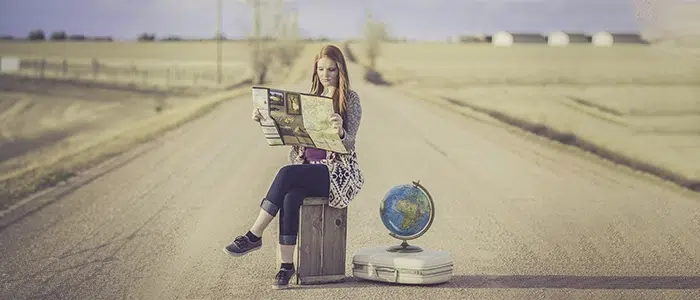 Mujer sentada en el medio de una carretera con su valija, mientras revisa un mapa.