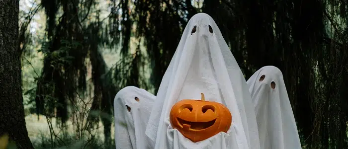Tres personas cubiertas con mantos blancos, simulando ser fantasmas, sosteniendo una calabaza en la mano.