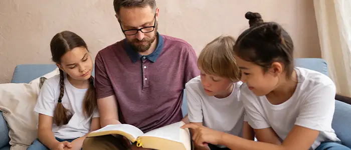 Hombre adulto leyendo un libro a tres niños de camisa blanca.