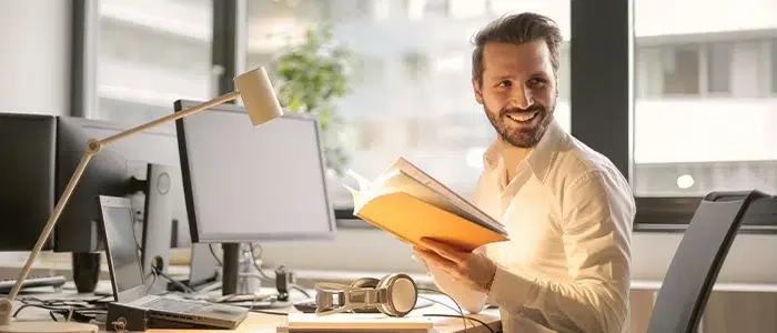 Persona con expresión de felicidad en su rostro, sentado frente a un computador sosteniendo un libro.